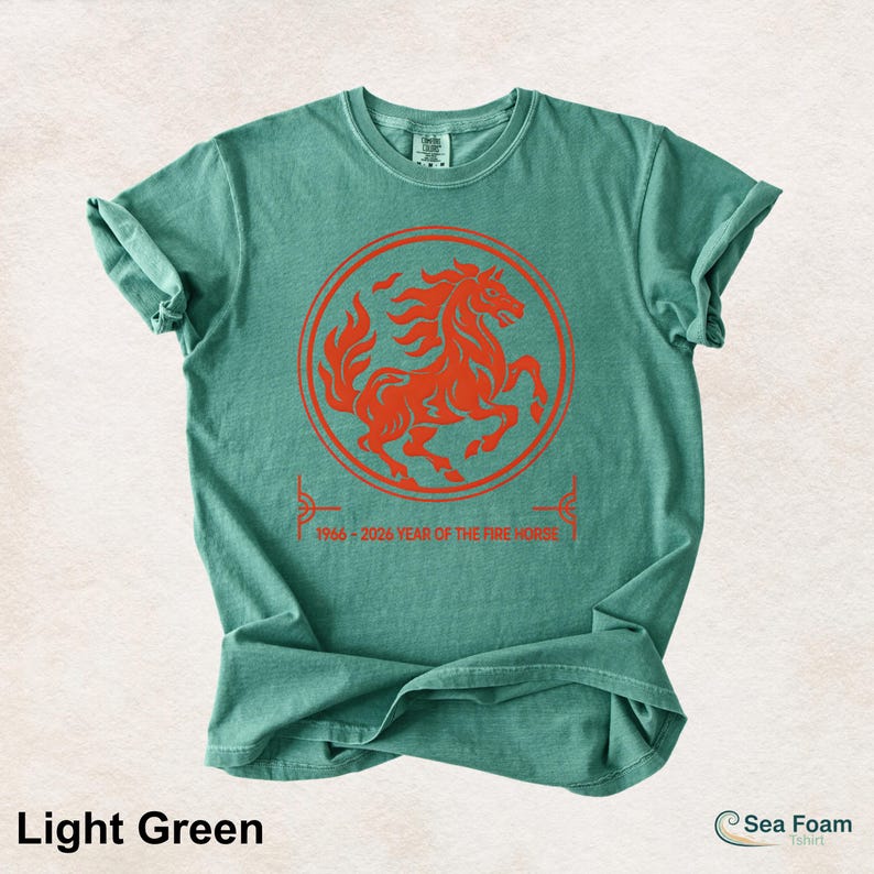 May include: A light green short-sleeve t-shirt featuring a circular orange graphic of a rearing horse. Below the circle, the text reads 1966 - 2026 YEAR OF THE FIRE HORSE. The shirt has a crew neckline and a standard fit with a soft, washed texture.