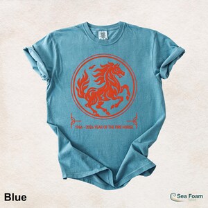 May include: A blue short-sleeve t-shirt featuring a red circular graphic of a rearing horse. Below the circle, the text reads 1966-2026 YEAR OF THE FIRE HORSE. The shirt has a crew neckline and a casual, relaxed fit with rolled sleeves.