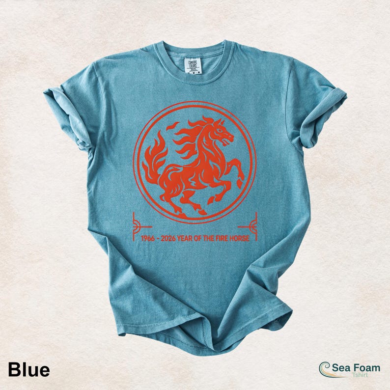 May include: A blue short-sleeve t-shirt featuring a red circular graphic of a rearing horse. Below the circle, the text reads 1966-2026 YEAR OF THE FIRE HORSE. The shirt has a crew neckline and a casual, relaxed fit with rolled sleeves.