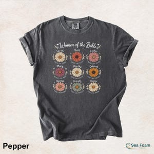 May include: A dark gray t-shirt featuring the text "Women of the Bible" above nine daisy illustrations, each with a woman's name and a related phrase. The shirt is a Pepper color.