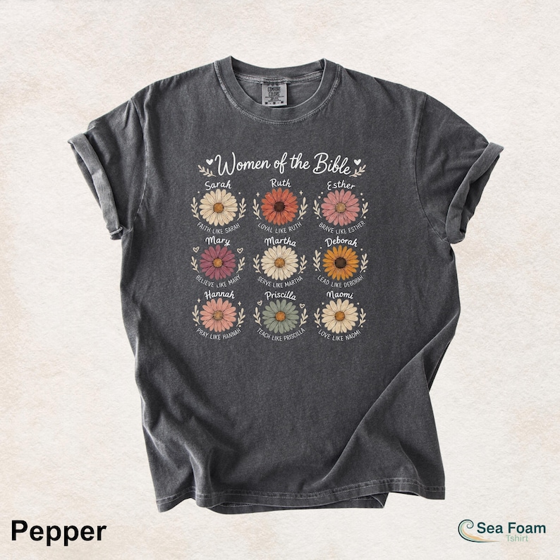 May include: A dark gray t-shirt featuring the text "Women of the Bible" above nine daisy illustrations, each with a woman's name and a related phrase. The shirt is a Pepper color.