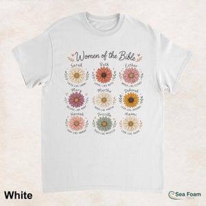 May include: White t-shirt featuring the text "Women of the Bible" above floral illustrations with names like Sarah, Ruth, and Esther. Each flower has a corresponding inspirational phrase. The shirt is made of a soft material.