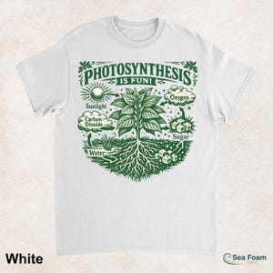 May include: White t-shirt with a green graphic illustrating photosynthesis. The design includes a tree, sunlight, carbon dioxide, water, oxygen, and sugar, with the text "PHOTOSYNTHESIS IS FUN!" The shirt has a crew neck and short sleeves.