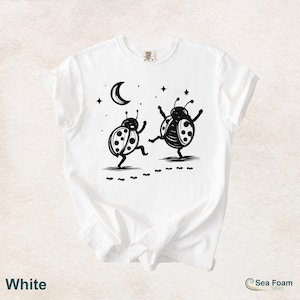 May include: White t-shirt featuring a black and white illustration of two dancing ladybugs under a crescent moon and stars. The shirt has a crew neck and short sleeves. The word "White" is printed at the bottom left.