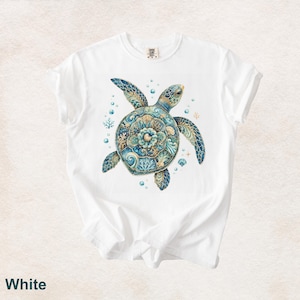 Puede incluir: Una camiseta blanca de manga corta con un gr&aacute;fico central de una tortuga marina con un caparaz&oacute;n decorado con intrincados patrones florales y de coral en azul y dorado, rodeada de peque&ntilde;as burbujas, ramas de coral y conchas marinas.