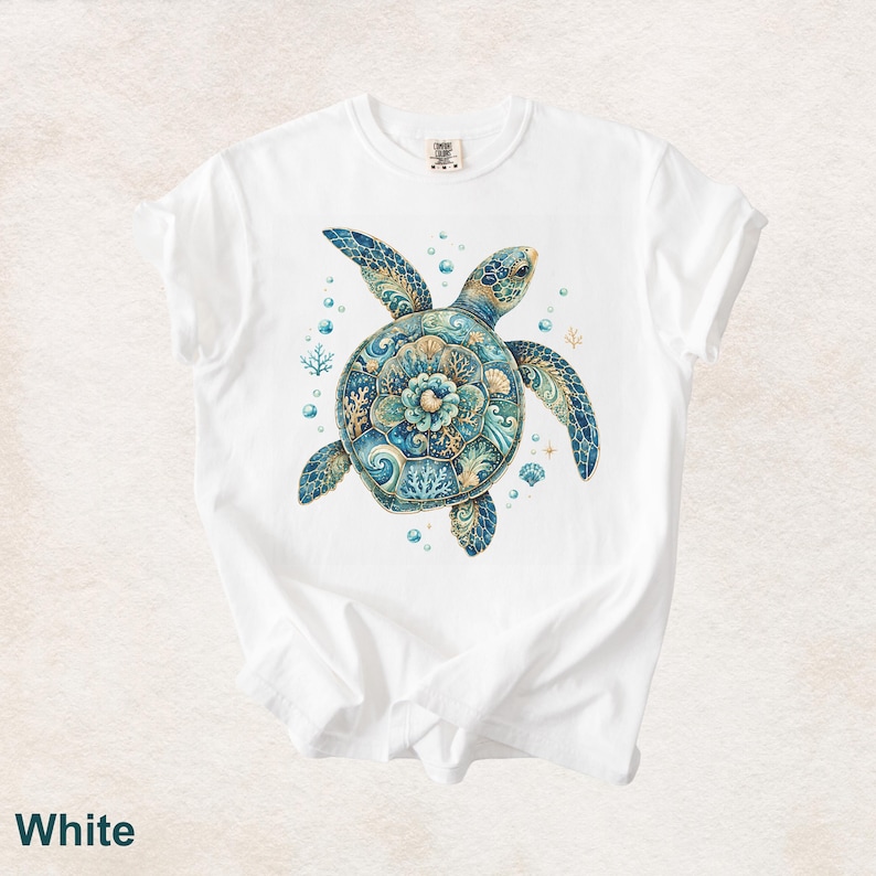 Puede incluir: Una camiseta blanca de manga corta con un gr&aacute;fico central de una tortuga marina con un caparaz&oacute;n decorado con intrincados patrones florales y de coral en azul y dorado, rodeada de peque&ntilde;as burbujas, ramas de coral y conchas marinas.