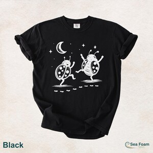May include: Black t-shirt featuring a whimsical design of two dancing ladybugs under a crescent moon and stars. The ladybugs are white with black spots, and the shirt has the text "Sea Foam t-shirt".