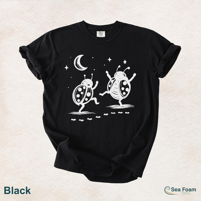 May include: Black t-shirt featuring a whimsical design of two dancing ladybugs under a crescent moon and stars. The ladybugs are white with black spots, and the shirt has the text "Sea Foam t-shirt".