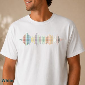 May include: A white short-sleeved t-shirt featuring a central graphic design composed of multicolored dots arranged in a horizontal sound wave pattern. The dots transition through various muted shades including blue, yellow, green, and pink.