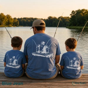 May include: Blue jean t-shirts with a white graphic of a person and two children fishing on a dock. The t-shirts are blue and are being worn by a person and two children. The image is taken outdoors near a lake.