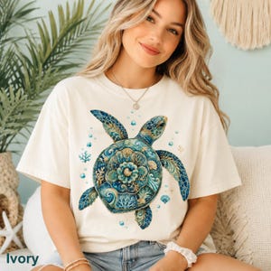 Puede incluir: Una camiseta de manga corta color marfil con un gr&aacute;fico grande y detallado de una tortuga marina. El caparaz&oacute;n de la tortuga presenta intrincados patrones en azul, turquesa y dorado, rodeado de peque&ntilde;as burbujas, corales y conchas marinas.