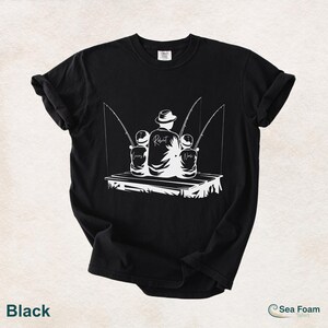 May include: Black t-shirt with a white graphic of a father and two children fishing on a dock. The father's back is turned, and the children's names, Lucas and Noah, are printed on the shirt. The word "Robert" is printed on the father's back.