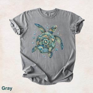 Puede incluir: Una camiseta gris de manga corta con un gr&aacute;fico central de una tortuga marina. El caparaz&oacute;n de la tortuga est&aacute; decorado con intrincados patrones azules, turquesas y dorados que recuerdan al coral y las olas del mar, rodeado de peque&ntilde;as burbujas.