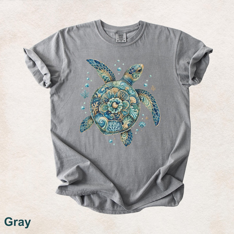 Puede incluir: Una camiseta gris de manga corta con un gr&aacute;fico central de una tortuga marina. El caparaz&oacute;n de la tortuga est&aacute; decorado con intrincados patrones azules, turquesas y dorados que recuerdan al coral y las olas del mar, rodeado de peque&ntilde;as burbujas.