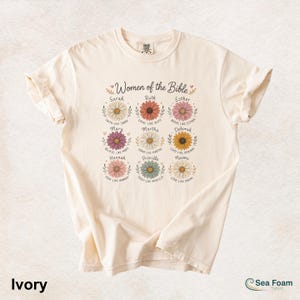 May include: Ivory-colored t-shirt featuring the text "Women of the Bible" above colorful daisy illustrations. Each daisy has a name and a related phrase. The shirt also has the text "Ivory" and the brand name "Sea Foam".