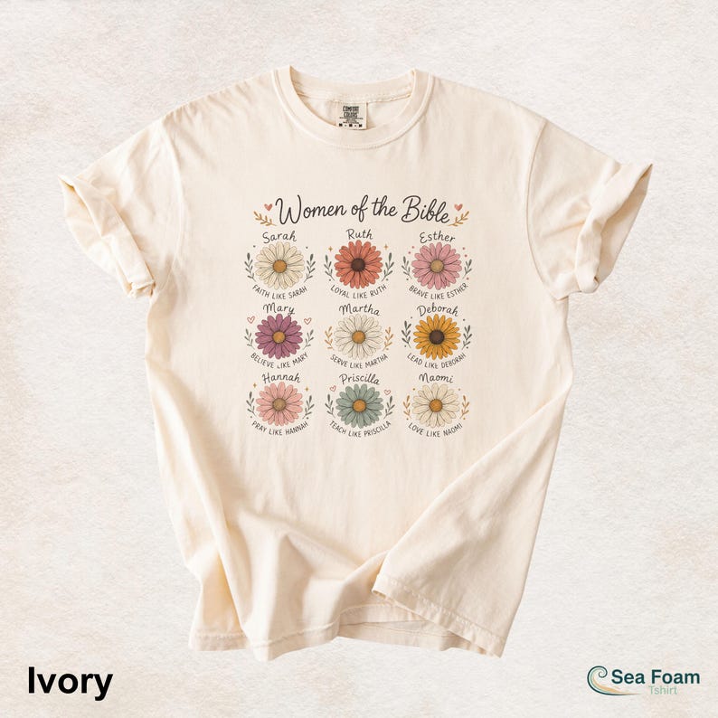 May include: Ivory-colored t-shirt featuring the text "Women of the Bible" above colorful daisy illustrations. Each daisy has a name and a related phrase. The shirt also has the text "Ivory" and the brand name "Sea Foam".