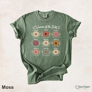 May include: Moss green t-shirt featuring the text "Women of the Bible" above nine daisy illustrations, each with a woman's name and a related phrase. The shirt has rolled-up sleeves and a crew neck.