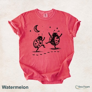 May include: Watermelon-colored t-shirt featuring a whimsical design of two dancing ladybugs under a crescent moon and stars. The ladybugs are black, and the shirt has a relaxed fit. The text "Watermelon" is at the bottom.