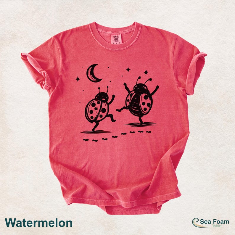 May include: Watermelon-colored t-shirt featuring a whimsical design of two dancing ladybugs under a crescent moon and stars. The ladybugs are black, and the shirt has a relaxed fit. The text "Watermelon" is at the bottom.