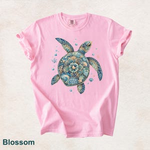 Puede incluir: Una camiseta de manga corta de color rosa claro con un gr&aacute;fico central de una tortuga marina con un caparaz&oacute;n decorado en intrincados patrones azules, turquesas y dorados de coral, olas y conchas, rodeada de peque&ntilde;as burbujas y estrellas.