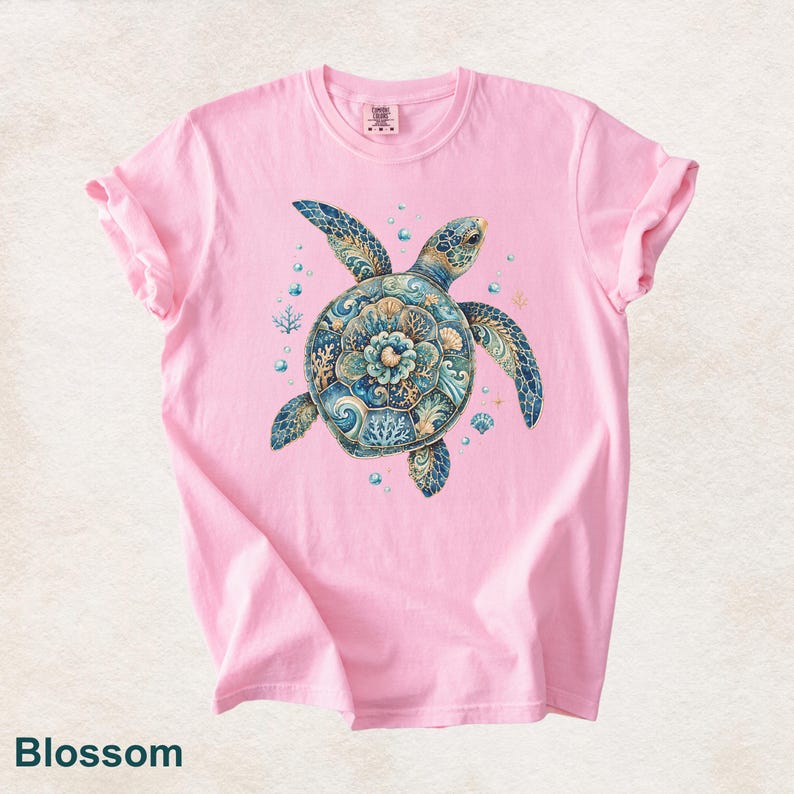 Puede incluir: Una camiseta de manga corta de color rosa claro con un gr&aacute;fico central de una tortuga marina con un caparaz&oacute;n decorado en intrincados patrones azules, turquesas y dorados de coral, olas y conchas, rodeada de peque&ntilde;as burbujas y estrellas.