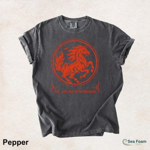 May include: A charcoal gray short-sleeve t-shirt featuring a vibrant orange graphic of a horse inside a circular border. Below the circle, the text reads 1966 2026 YEAR OF THE FIRE HORSE. The shirt has a crew neckline and a washed-out fabric texture.