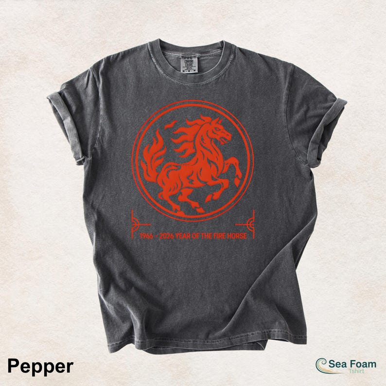 May include: A charcoal gray short-sleeve t-shirt featuring a vibrant orange graphic of a horse inside a circular border. Below the circle, the text reads 1966 2026 YEAR OF THE FIRE HORSE. The shirt has a crew neckline and a washed-out fabric texture.