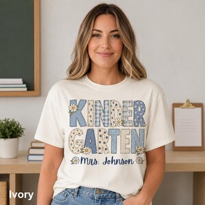 Może przedstawiać: T-shirt z krótkim rękawem w kolorze kości słoniowej z napisem KINDERGARTEN dużymi, blokowymi literami z niebiesko-beżowym wzorem patchworkowym w kratkę vichy, kwiaty i kropki. Poniżej znajduje się napis Mrs. Johnson wykonany niebieską czcionką.