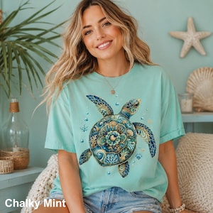 Puede incluir: Una camiseta de manga corta en color verde menta tiza con un gr&aacute;fico grande y detallado de una tortuga marina. El caparaz&oacute;n de la tortuga est&aacute; decorado con intrincados patrones florales y de coral en azul, turquesa y oro, rodeado de peque&ntilde;as burbujas.