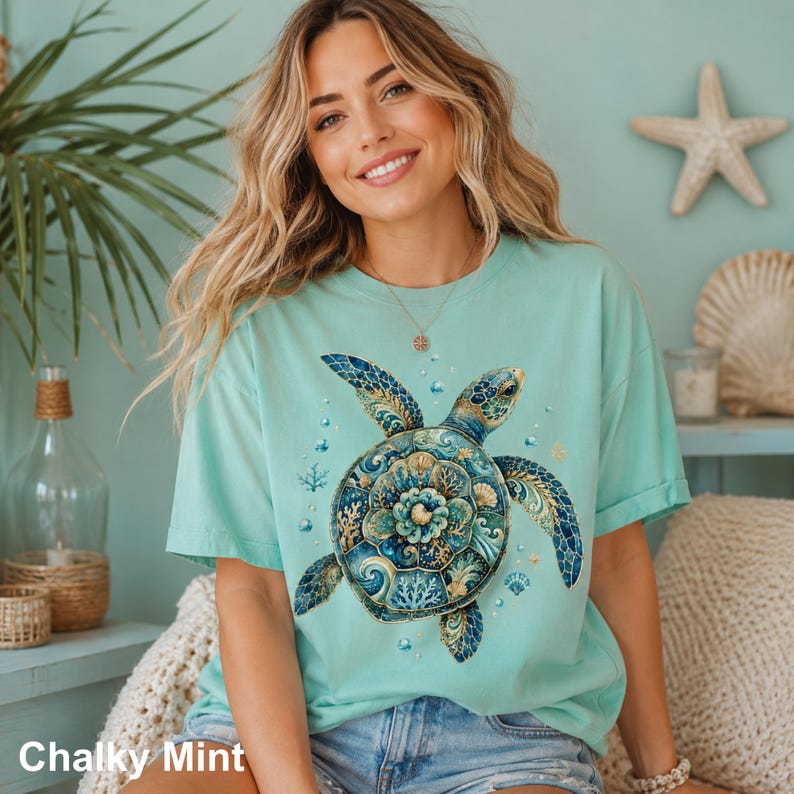 Puede incluir: Una camiseta de manga corta en color verde menta tiza con un gr&aacute;fico grande y detallado de una tortuga marina. El caparaz&oacute;n de la tortuga est&aacute; decorado con intrincados patrones florales y de coral en azul, turquesa y oro, rodeado de peque&ntilde;as burbujas.