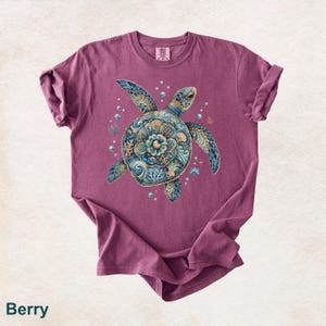 Puede incluir: Una camiseta de manga corta color baya con un gr&aacute;fico central de una tortuga marina con un caparaz&oacute;n estampado en tonos azules, verde azulado y dorados. La tortuga est&aacute; rodeada de peque&ntilde;as burbujas, corales y conchas. Cuello redondo.