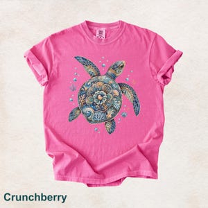 Puede incluir: Una camiseta de manga corta de color rosa brillante con un gr&aacute;fico central de una tortuga marina con un patr&oacute;n de caparaz&oacute;n detallado en tonos azul, dorado y blanco. La tortuga est&aacute; rodeada de peque&ntilde;as burbujas y detalles de coral.