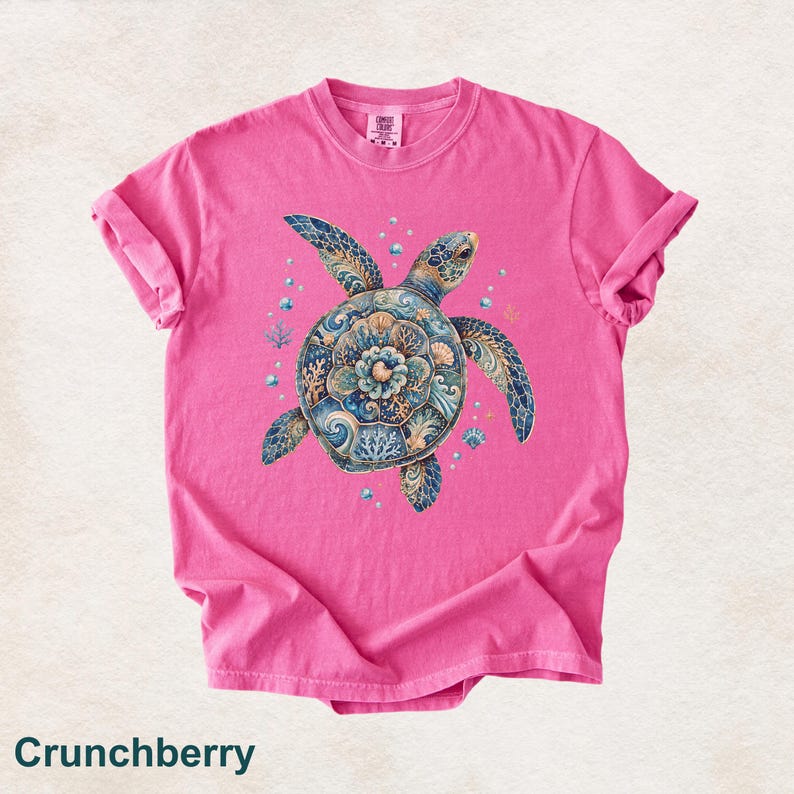 Puede incluir: Una camiseta de manga corta de color rosa brillante con un gr&aacute;fico central de una tortuga marina con un patr&oacute;n de caparaz&oacute;n detallado en tonos azul, dorado y blanco. La tortuga est&aacute; rodeada de peque&ntilde;as burbujas y detalles de coral.