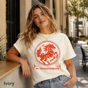 May include: An ivory short-sleeve t-shirt featuring a red circular graphic of a stylized horse. Below the circle, the text reads 1966 - 2026 YEAR OF THE FIRE HORSE. The design is centered on the chest of the garment, which has a crew neckline.
