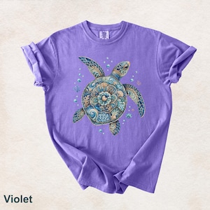 Puede incluir: Una camiseta violeta de manga corta con un gr&aacute;fico central de una tortuga marina. El caparaz&oacute;n de la tortuga est&aacute; decorado con intrincados patrones azules, turquesas y dorados que recuerdan al coral y a las olas del mar.