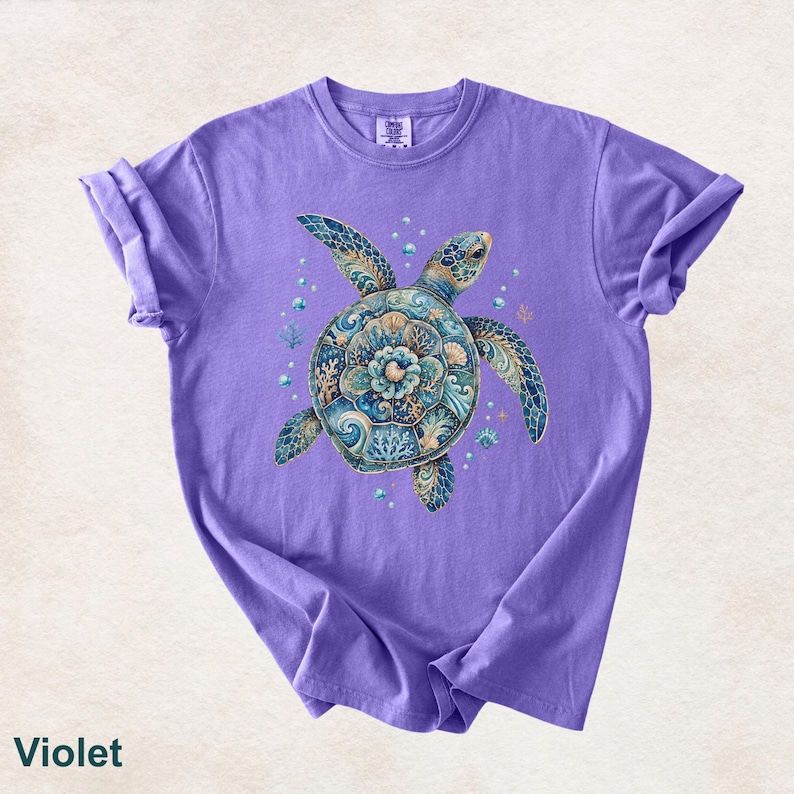 Puede incluir: Una camiseta violeta de manga corta con un gr&aacute;fico central de una tortuga marina. El caparaz&oacute;n de la tortuga est&aacute; decorado con intrincados patrones azules, turquesas y dorados que recuerdan al coral y a las olas del mar.