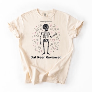 Può includere: T-shirt color crema con illustrazione di uno scheletro nero. Il testo "Undiagnosed" è sopra e "But Peer Reviewed" sotto. Lo scheletro fa il pollice in su, circondato da scarabocchi di tazze di caffè, cuori e fulmini.