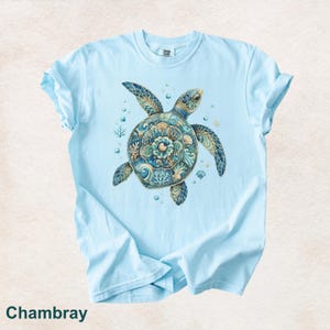 Puede incluir: Una camiseta de manga corta de color azul claro con un gr&aacute;fico central de una tortuga marina con un caparaz&oacute;n decorado con intrincados patrones en azul, turquesa y dorado, rodeada de peque&ntilde;as burbujas y elementos de coral.