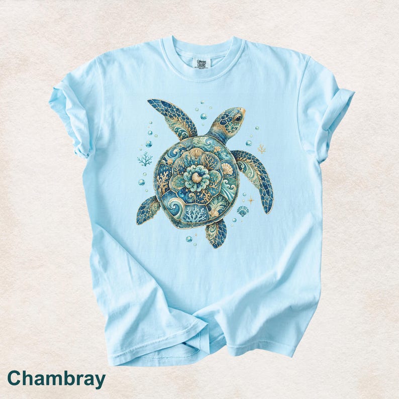 Puede incluir: Una camiseta de manga corta de color azul claro con un gr&aacute;fico central de una tortuga marina con un caparaz&oacute;n decorado con intrincados patrones en azul, turquesa y dorado, rodeada de peque&ntilde;as burbujas y elementos de coral.