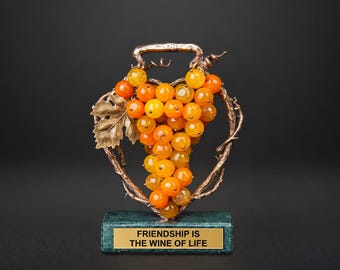 Peut inclure: Une sculpture décorative composée d'une grappe de perles sphériques translucides couleur ambre, entourée d'un cadre en métal bronze en forme de vigne. Elle repose sur un socle rectangulaire en marbre vert avec une plaque dorée portant l'inscription FRIENDSHIP IS THE WINE OF LIFE.