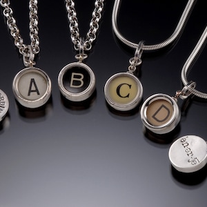 May include: Five silver necklaces with typewriter key pendants. The keys spell out "EXTRAORDINARY" and "A, B, C, D".