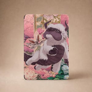May include: A rectangular tablet case featuring a white, furry, six-legged creature with curved horns and a brown belly, flying alongside a small winged creature. The background displays pink clouds, green leaves, and white lotus flowers.