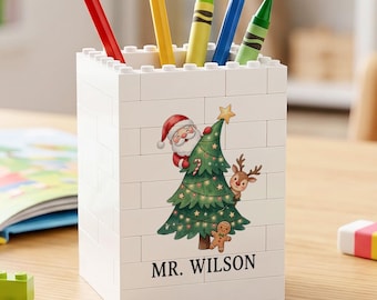 May include: A white rectangular pencil holder constructed from interlocking plastic building blocks. The front features a printed illustration of a Christmas tree with Santa Claus, a reindeer, and a gingerbread man, with the text MR. WILSON below.