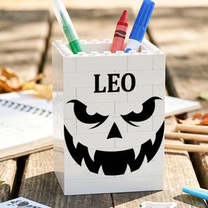 May include: A white rectangular pencil holder constructed from interlocking plastic building blocks. The front features the name LEO printed in black above a black graphic of a jagged, toothy monster face. Three writing utensils are inside.