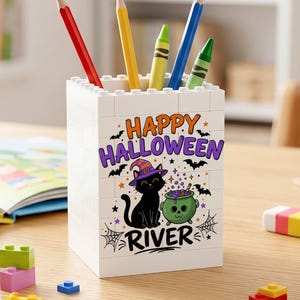 May include: A white rectangular pencil holder constructed from interlocking building blocks. The front features a graphic of a black cat wearing a purple witch hat next to a green skull cauldron, with the text Happy Halloween and the name River.