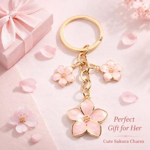 May include: A gold-colored keychain with three pink flower charms and a larger pink flower charm at the bottom. The keychain is on a pink surface with a pink gift box and scattered pink flower petals.
