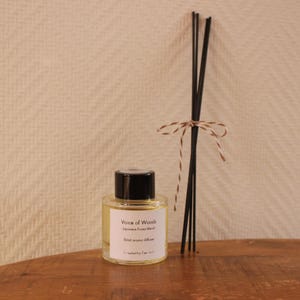 May include: Aroma diffuser featuring a clear glass bottle with a black cap, filled with a pale yellow liquid. The label reads "Voice of Woods Japanese Forest Blend 50ml aroma diffuser." Black reeds are tied with twine. The items are on a wooden surface.