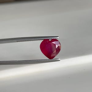 Op de afbeelding: Een hartvormige, gefacetteerde robijn edelsteen, vastgehouden door een zilveren pincet. De robijn heeft een diepe, rijke rode kleur, met meerdere facetten die het licht vangen. De pincet is scherp gefocust.