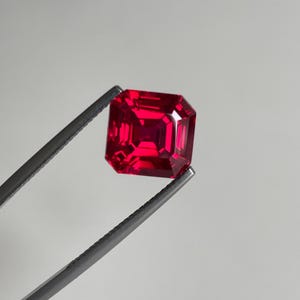 May include: A vibrant, deep red gemstone with a rectangular emerald cut, featuring sharp, clean facets that reflect light. The stone is held by a pair of dark grey metal tweezers, showcasing its saturated color and precise geometric shape.