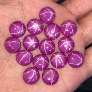May include: A collection of round, polished star ruby gemstones. Each gem is a deep magenta colour with a six-rayed star pattern across its surface. The gems are displayed against a neutral background.